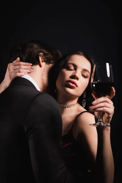Mulher sensual com olhos fechados segurando copo de vinho tinto, enquanto o homem de terno preto abraçando-a isolada no preto — Stock Photo