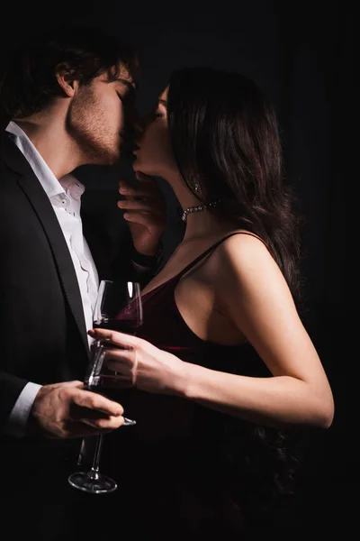 Jovem casal elegante segurando copos de vinho enquanto se beija com os olhos fechados à noite — Fotografia de Stock