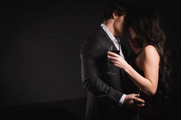 Élégant jeune couple avec des verres de vin rouge embrassant à la maison dans l'obscurité — Photo de stock