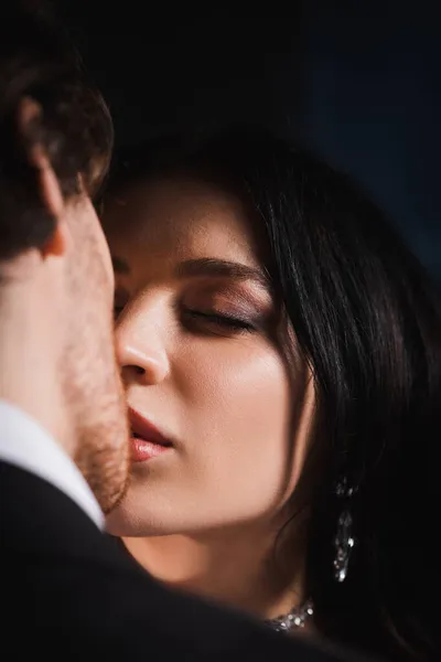 Vue rapprochée de jeune femme brune aux yeux fermés embrassant homme flou sur fond sombre — Photo de stock