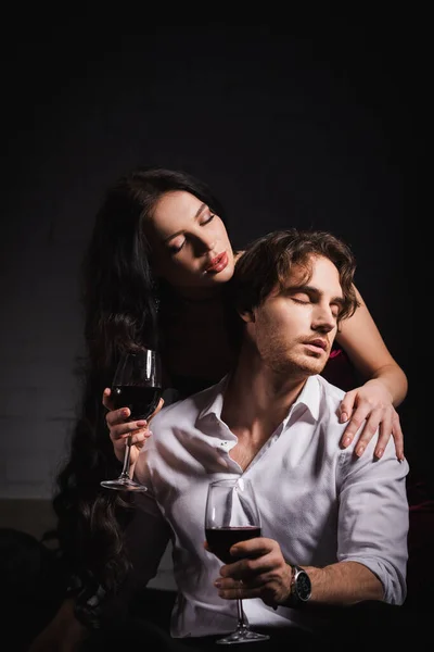 Donna appassionata con bicchiere di rosso mentre seduce giovane in camicia bianca in camera da letto scura — Foto stock