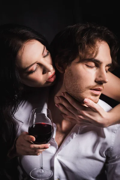 Passionate woman with glass of red whine embracing man in white shirt on dark background — Stock Photo