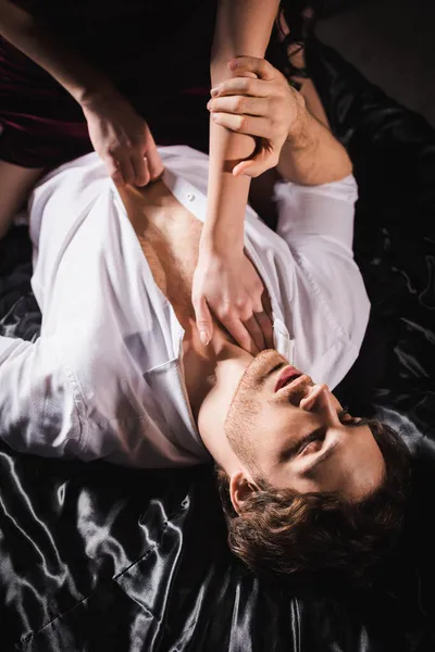 Alto angolo di vista della donna spogliarsi uomo in camicia bianca sdraiato con gli occhi chiusi su biancheria da letto in raso nero — Foto stock