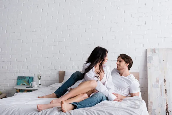 Seductive woman in shirt and bra seducing man lying on bed in jeans — Stock Photo