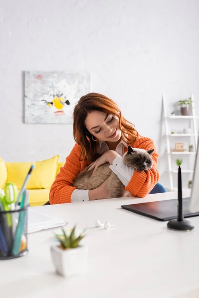 Счастливая женщина, обнимающая кота, сидя за столом рядом с размытым графическим планшетом и стилусом — стоковое фото