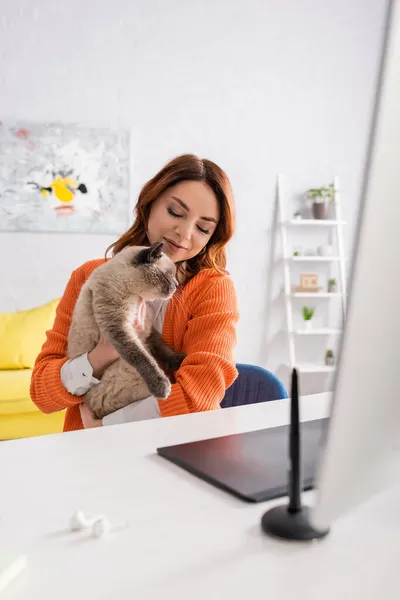 Femme heureuse assise avec chat près de tablette graphique floue et stylet sur le bureau à la maison — Photo de stock