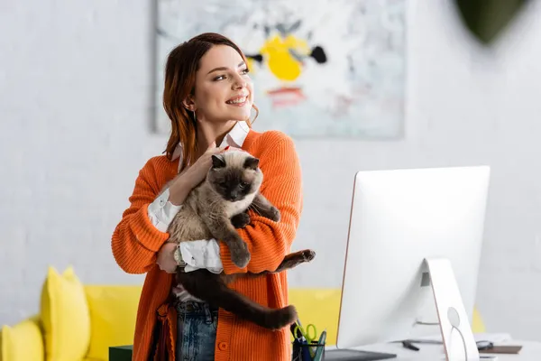 Cheerful young woman looking away while standing with cat near computer monitor — Stock Photo