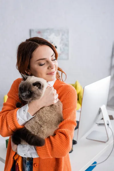 Glückliche Frau mit geschlossenen Augen, die Katze in der Nähe des Computermonitors auf verschwommenem Hintergrund umarmt — Stockfoto