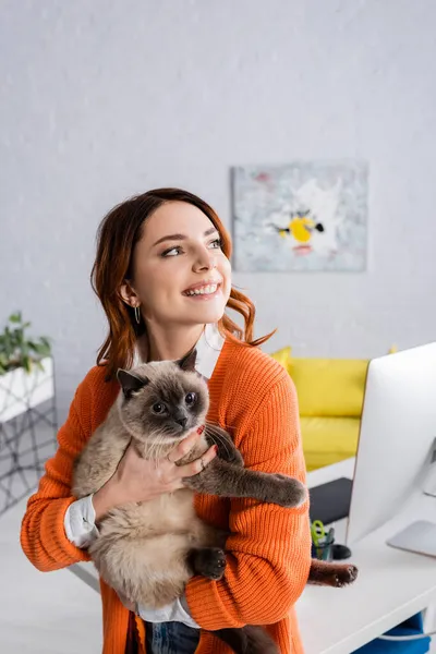 Mulher alegre segurando gato fofo enquanto está perto da mesa com monitor de computador em casa — Fotografia de Stock