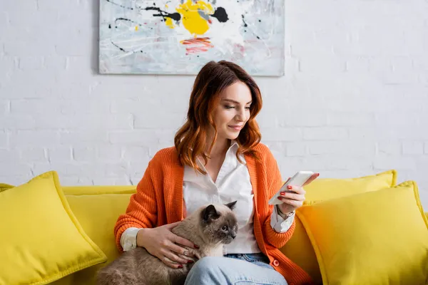 Lächelnde Frau, die auf dem Handy Nachrichten sendet, während sie mit Katze auf der gelben Couch sitzt — Stockfoto