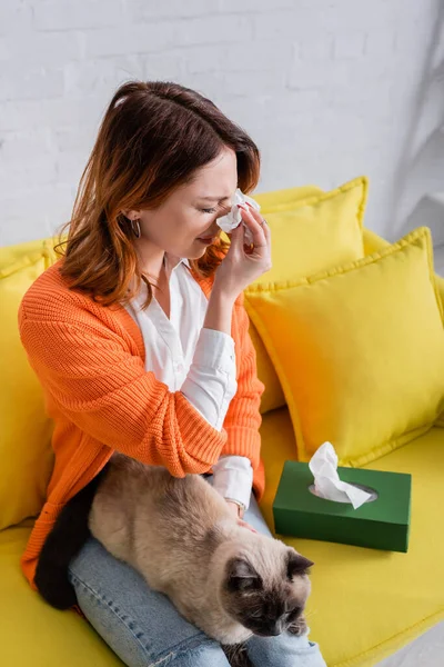 Junge Frau leidet an Allergie, während sie mit Katze auf dem Sofa sitzt — Stockfoto