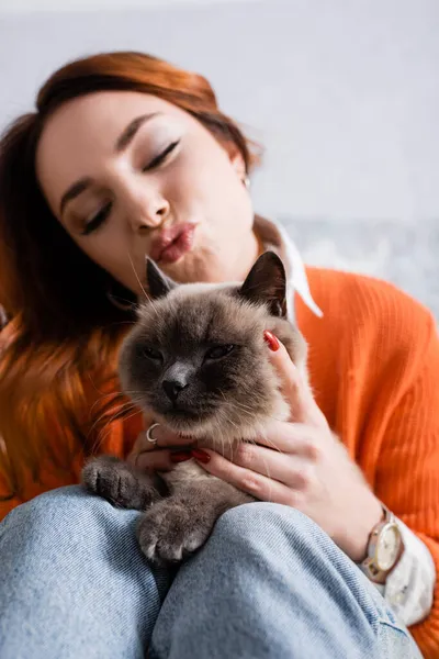 Femme floue avec les yeux fermés boudant lèvres près du chat pelucheux à la maison — Photo de stock