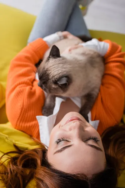 Vista superior da mulher sorridente com os olhos fechados sentados com gato fofo no sofá em casa — Fotografia de Stock