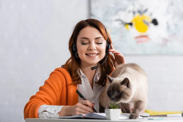 Mujer feliz en la escritura de auriculares en el cuaderno cerca del gato borroso en el escritorio - foto de stock