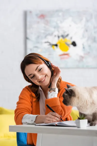 Mujer alegre en auriculares escritura en cuaderno mientras trabaja cerca del gato sentado en el escritorio - foto de stock