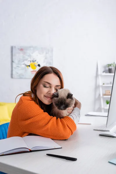 Donna felice abbracciare gatto mentre seduto vicino alla scrivania di lavoro a casa — Foto stock