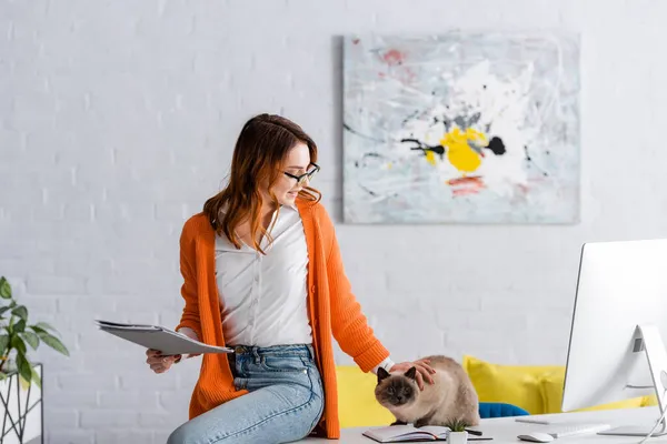 Jeune pigiste avec des documents caressant chat assis sur le bureau près de l'écran d'ordinateur — Photo de stock