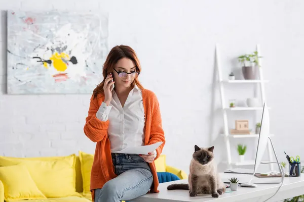 Young woman in eyeglasses holding documents and talking on smartphone while sitting on work desk near cat — Stock Photo