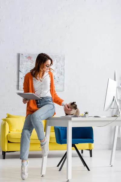 Femme heureuse avec des documents assis sur le bureau près du chat et moniteur d'ordinateur — Photo de stock