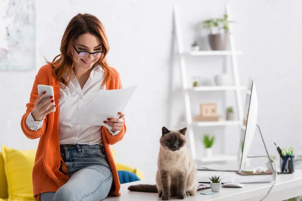 Cheerful freelancer with smartphone looking at documents near cat sitting on desk — Stock Photo