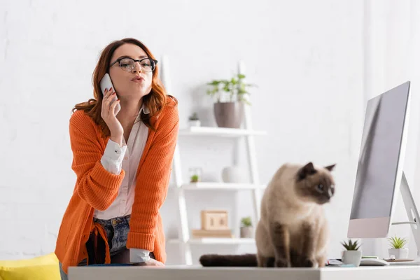 Femme en lunettes boudant les lèvres tout en appelant sur le téléphone portable près chat flou sur le bureau — Photo de stock