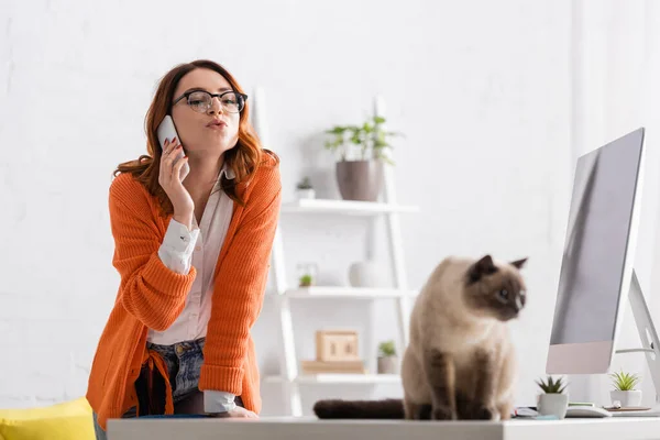 Woman in eyeglasses pouting lips while calling on cellphone near blurred cat on desk — Stock Photo