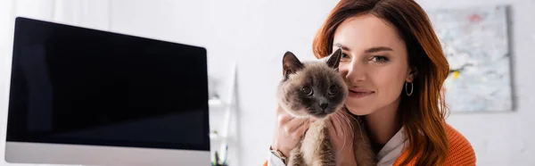 Mujer joven sonriendo a la cámara mientras sostiene gato cerca de monitor de computadora con pantalla en blanco, pancarta - foto de stock