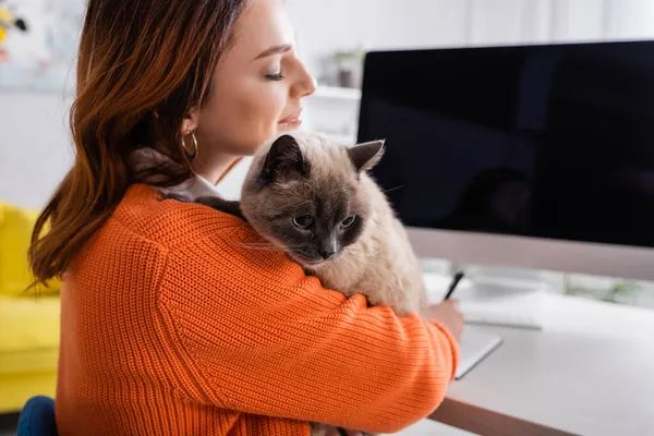 Mulher satisfeita com olhos fechados abraçando gato perto monitor borrado com tela em branco — Fotografia de Stock