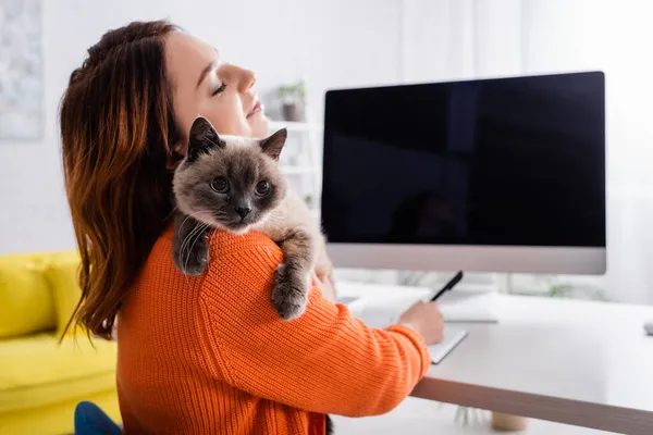 Mujer feliz con gato esponjoso en el hombro sentado cerca de monitor borroso con pantalla en blanco en casa - foto de stock