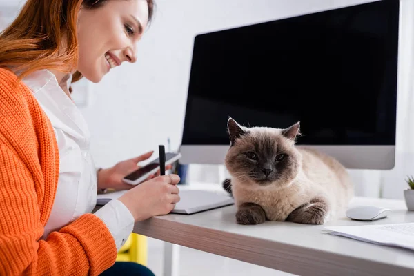 Donna sorridente con smartphone che scrive nel taccuino vicino al gatto sulla scrivania — Foto stock