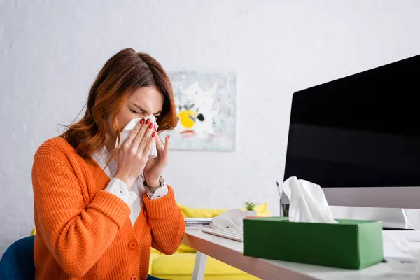 Mujer enferma con los ojos cerrados estornudos en servilleta de papel cerca del monitor de la computadora con pantalla en blanco - foto de stock