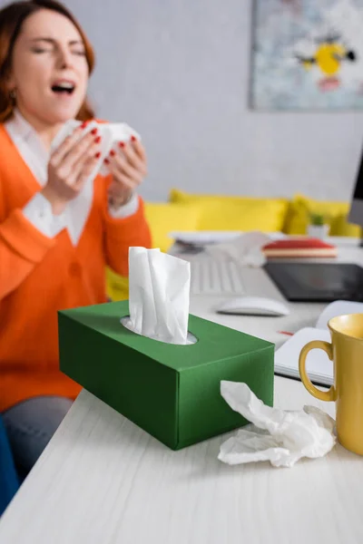 Selektiver Fokus der Packung mit Papierservietten in der Nähe einer niesenden kranken Frau auf verschwommenem Hintergrund — Stockfoto