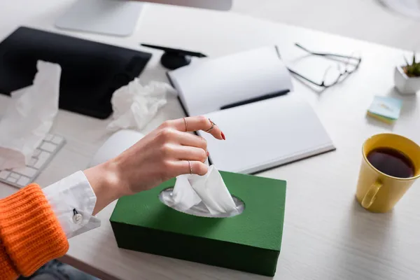 Ausgeschnittene Ansicht einer kranken Frau, die Papierservietten in der Nähe von leerem Notizbuch, Grafik-Tablet und Tee auf dem Schreibtisch nimmt — Stockfoto