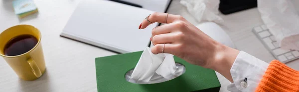 Abgeschnittene Ansicht einer Frau, die Papierserviette in der Nähe von Tasse Tee und Notizbuch auf dem Schreibtisch nimmt, Banner — Stockfoto