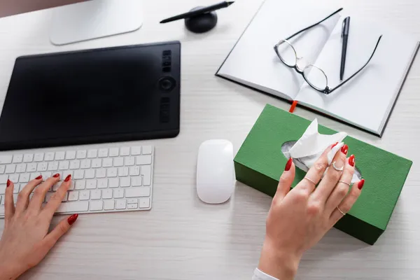 Vista superior de la mujer recortada tomando servilleta de papel del paquete cerca del teclado, ratón de la computadora, tableta gráfica y portátil - foto de stock