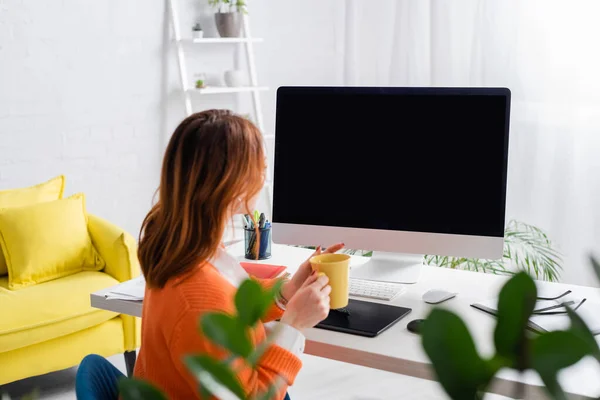 Retoque con taza de té cerca de la tableta gráfica y monitor con pantalla en blanco en casa - foto de stock