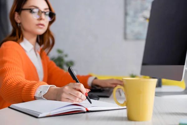 Blurred woman writing in notebook near cup of tea and computer monitor on desk — Stock Photo