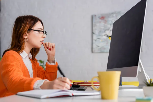 Thoughtful woman in eyeglasses writing in blurred notebook while looking at monitor with blank screen — Stock Photo