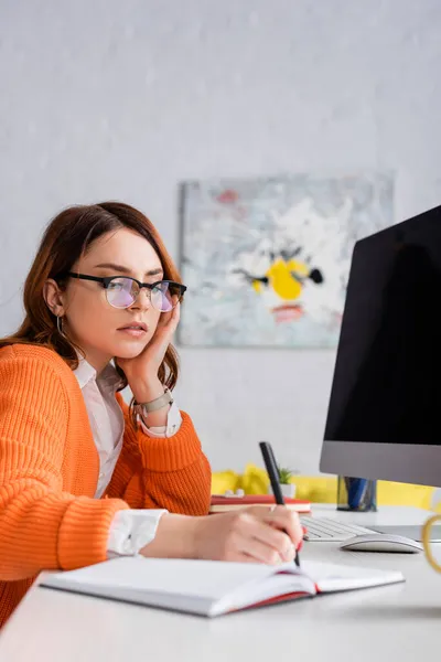 Frau mit Brille schreibt in verschwommenes Notizbuch neben Computermonitor mit leerem Bildschirm — Stockfoto