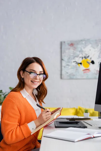 Happy retoucher in eyeglasses smiling at camera while writing in notebook near graphic tablet — Stock Photo