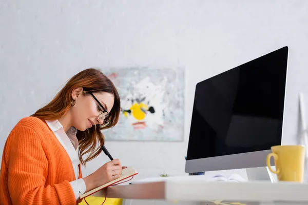 Freelancer in eyeglasses writing in notebook near computer monitor with blank screen — Stock Photo