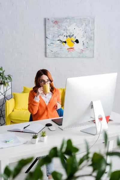 Joven diseñador en gafas tomando té mientras trabaja cerca del monitor de la computadora en casa - foto de stock