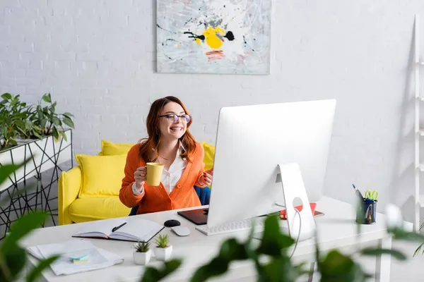 Designer feliz em óculos segurando xícara de chá enquanto sorri perto do monitor do computador — Fotografia de Stock