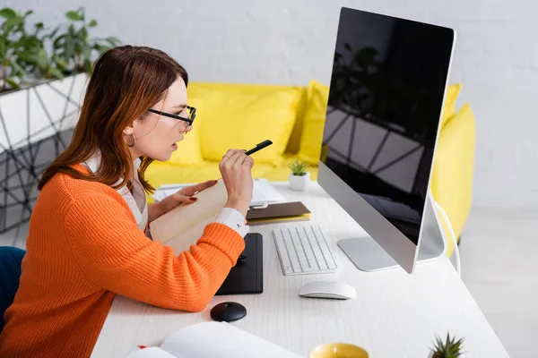 Freelancer in eyeglasses holding notebook and pointing with pen at monitor while working at home — Stock Photo