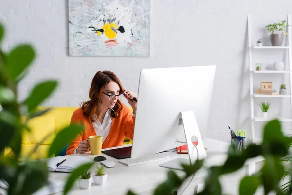 Concentrated retoucher adjusting eyeglasses while working near graphic tablet and monitor on blurred foreground — Stock Photo