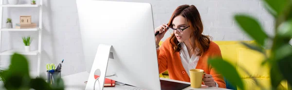 Woman with cup of tea touching eyeglasses while working near monitor on blurred foreground, banner — Stock Photo