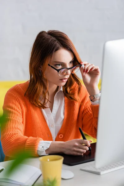 Concentrated freelancer touching eyeglasses while working on graphic tablet and looking at blurred monitor — Stock Photo