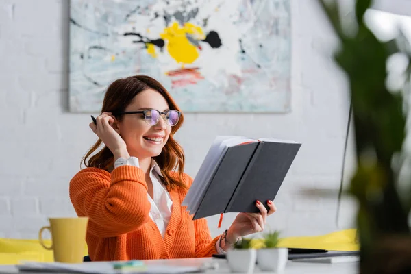 Cheerful woman in eyeglasses looking at notebook while working at home on blurred foreground — Stock Photo