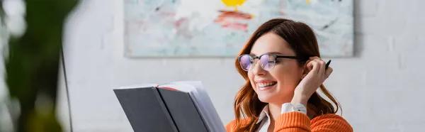 Donna gioiosa con gli occhiali che guarda il quaderno mentre lavora a casa, banner — Foto stock