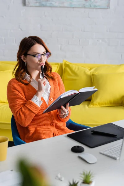 Thoughtful designer in eyeglasses looking in notebook near graphic tablet on desk — Stock Photo